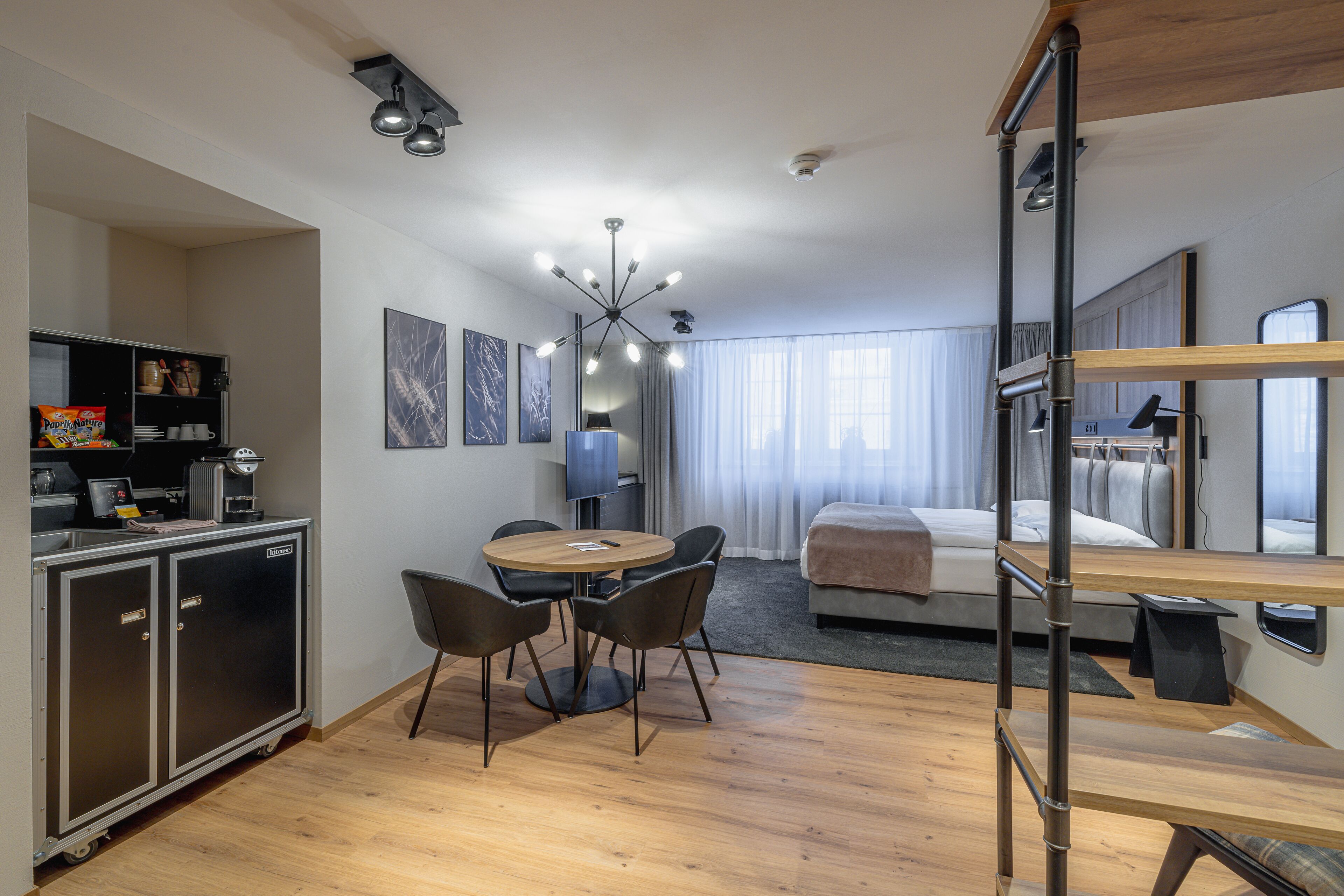 Habitación de hotel compacta con kitchenette, comedor con una mesa redonda y sillas, cama con cabecera y un ventanal con cortinas.