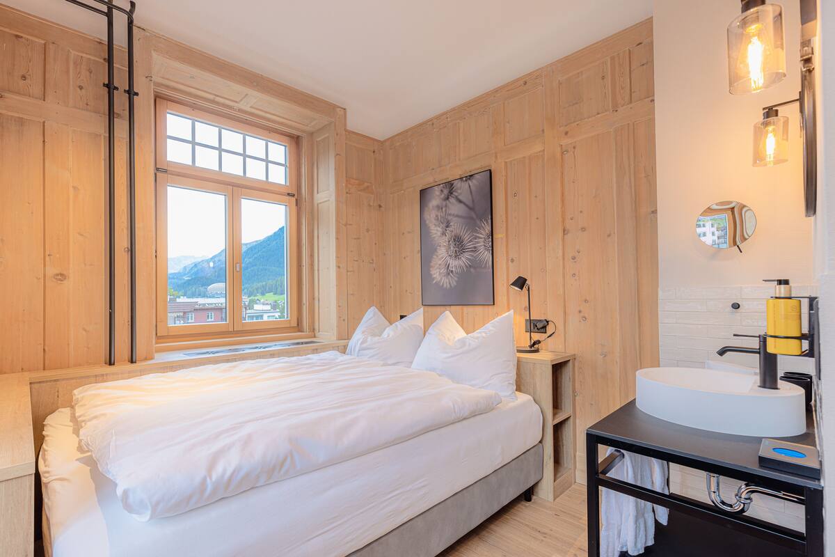 Un dormitorio con una cama grande, una ventana con vista a la montaña, una mesita de noche y un baño con lavabo y espejo.