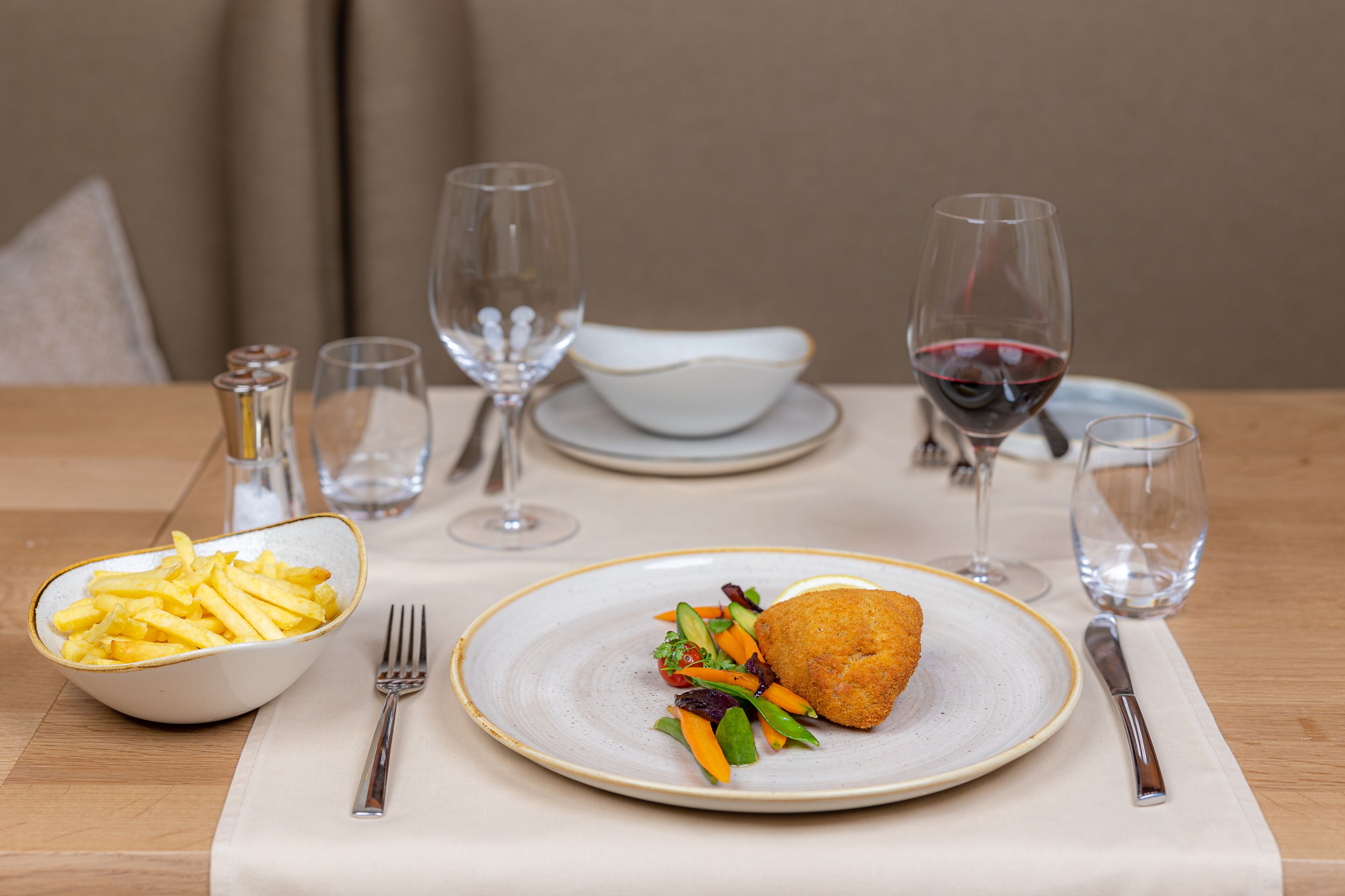 Una mesa de comedor con un plato de comida, un bol de papas fritas y dos copas de vino.