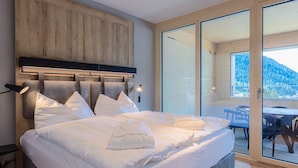 A bedroom with a large bed, a wooden headboard, and a sliding glass door leading to a balcony with a table and chairs. - ALPINE INN Davos (Davos)