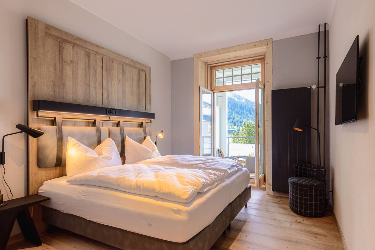 Un dormitorio con una cama grande, cabecera de madera, televisor montado en la pared y ventana con vistas al exterior.