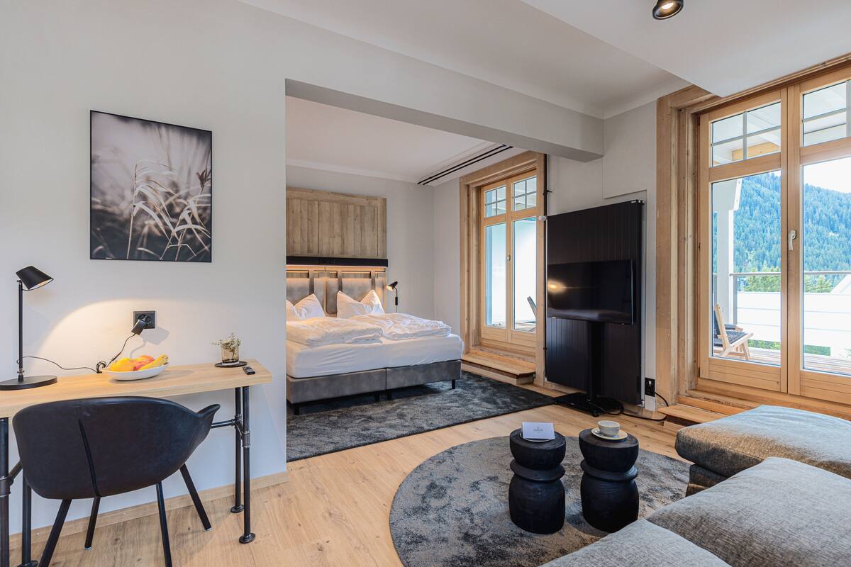 Un dormitorio moderno con cama, escritorio, silla, televisor y vistas al exterior.