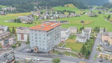 A multi-story building with a red roof, surrounded by a mix of residential and commercial buildings, set in a hilly area with green fields.