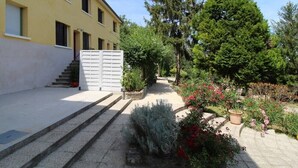 Un bâtiment à la façade jaune, doté d’une entrée fermée par un portail et d’un jardin où poussent différentes plantes et arbres.