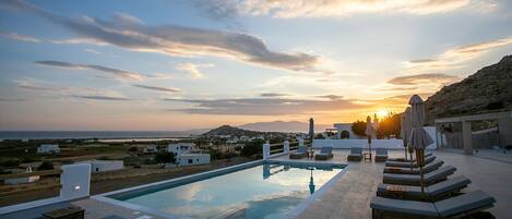 A rooftop pool with lounge chairs and umbrellas, overlooking a coastal landscape with buildings and mountains during sunset.