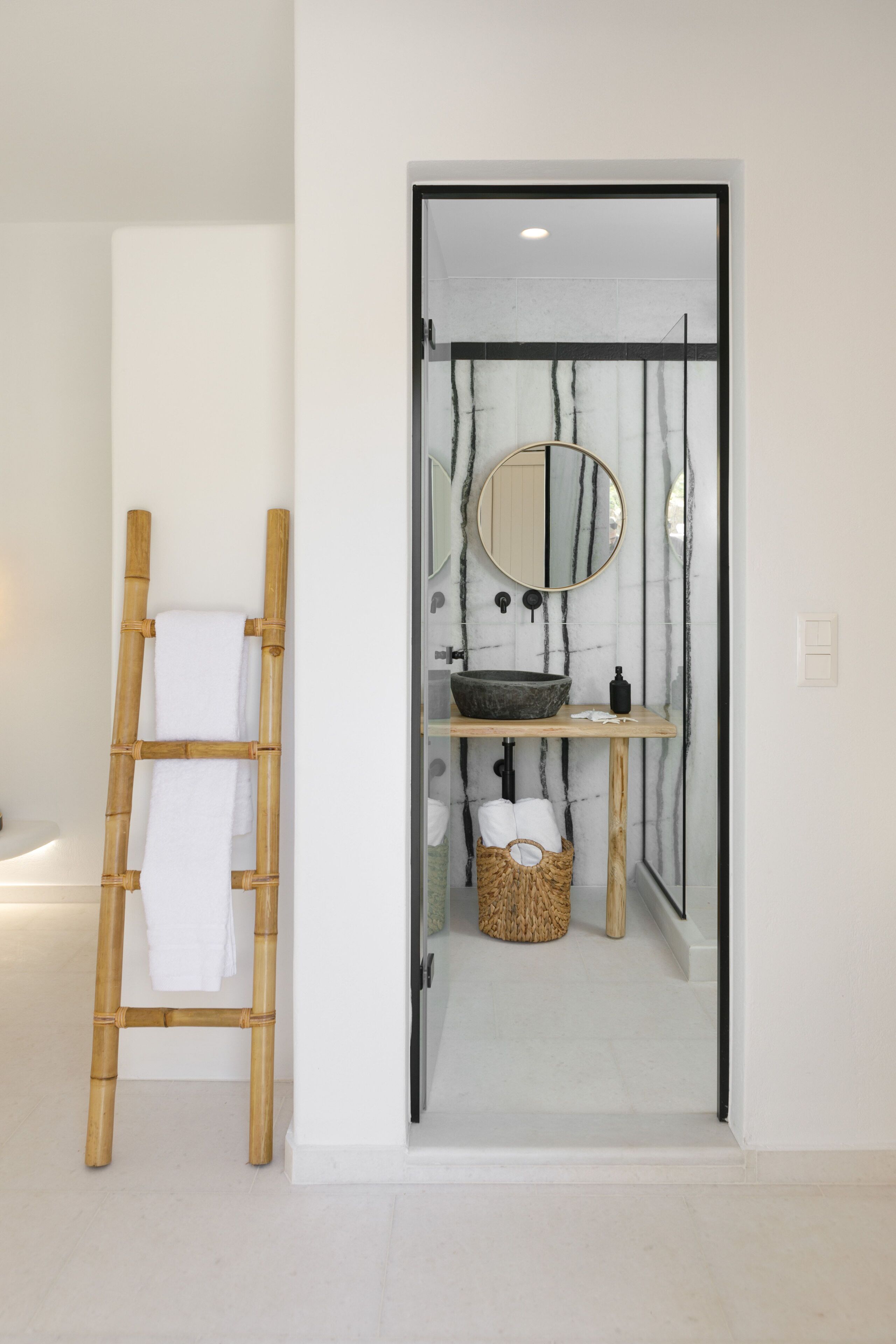 A bathroom with a wooden ladder, a round mirror, a stone basin, and a wicker basket.