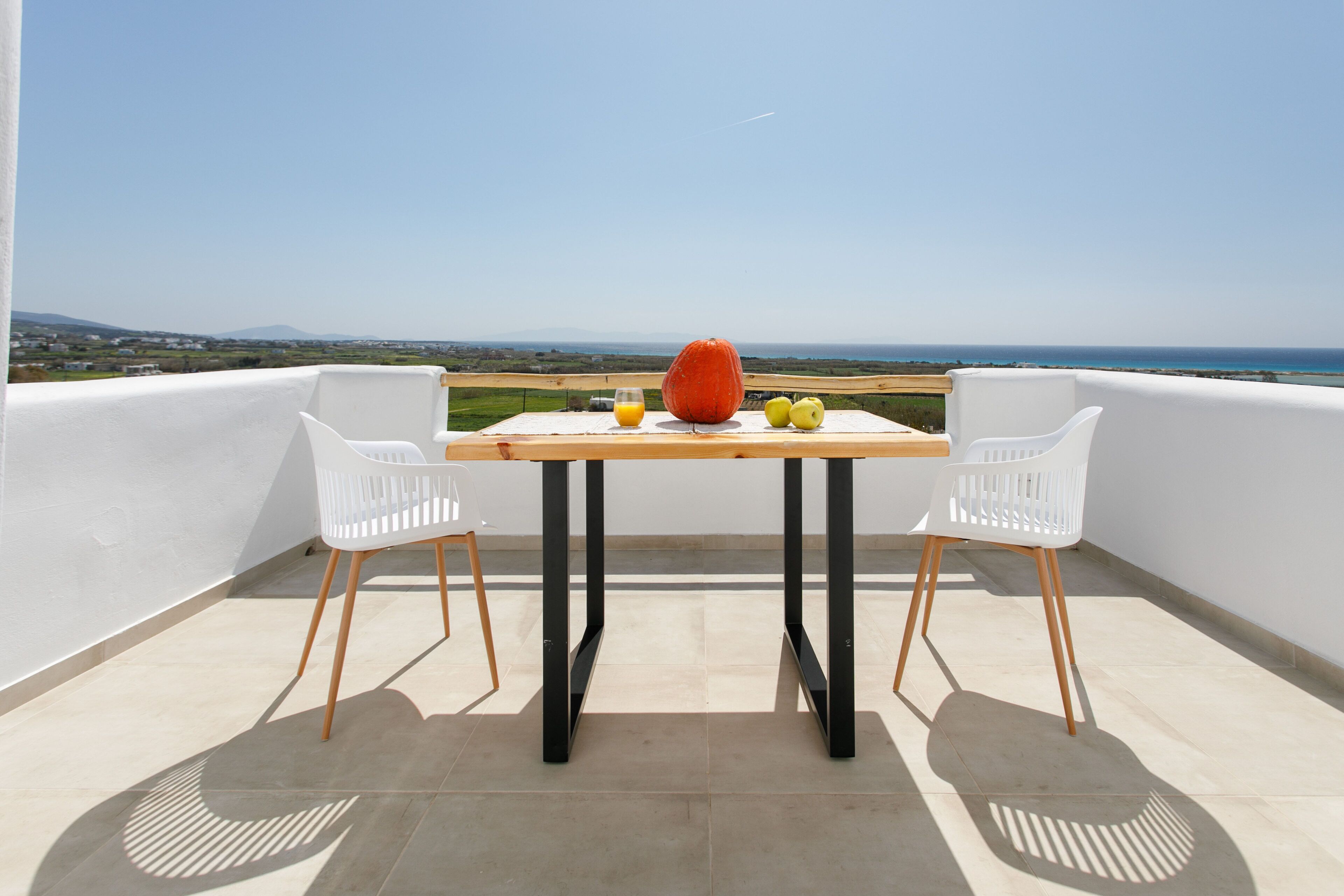 A balcony with a table, two white chairs, a wooden table, a glass of orange juice, a large orange fruit, and two green apples.