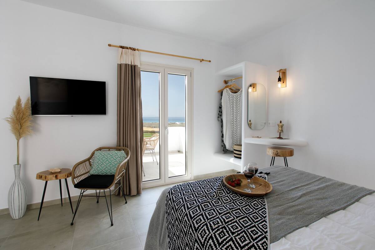 A bedroom with a bed, a TV, a chair, a small table, a tray with wine glasses, and a view through the window.