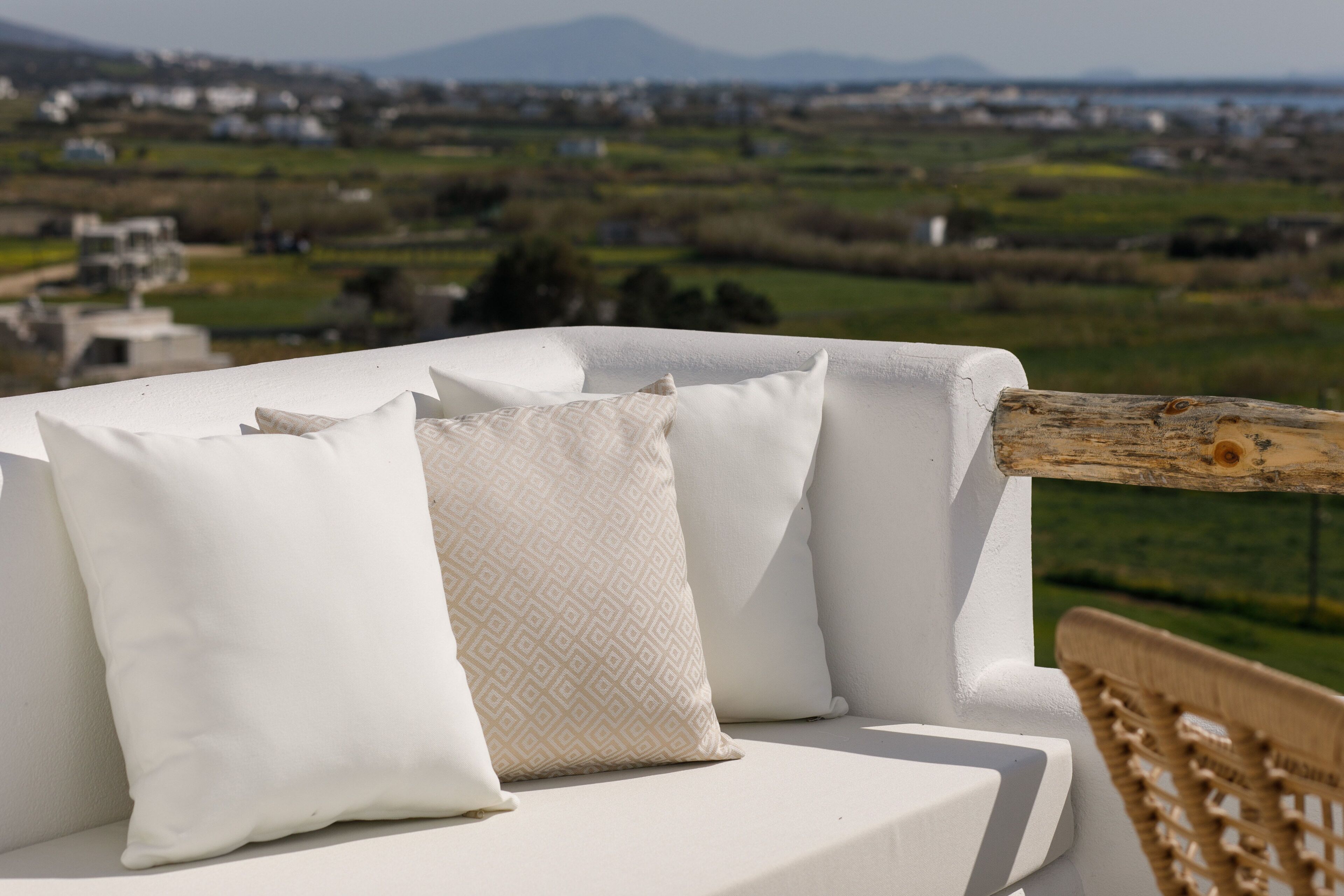 Un canapé d’extérieur blanc, agrémenté de coussins à motifs, offrant une vue imprenable sur un paysage pittoresque, avec des maisons et de la verdure.