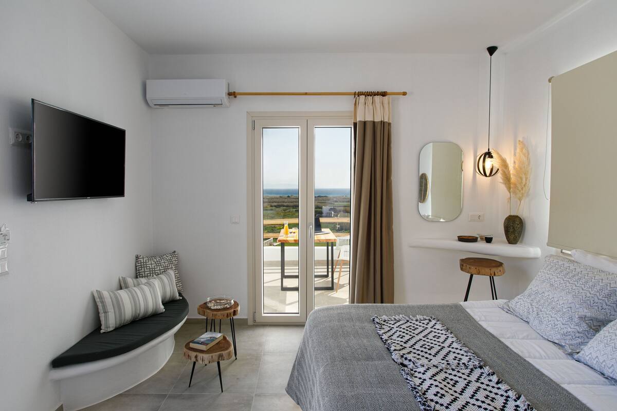 A modern bedroom with a bed, a TV, a balcony with a table and chairs, and a view of the landscape.