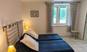 A room with two beds, a window with curtains, a small stool, and a wall-mounted picture.