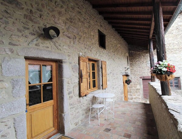 Un edificio de piedra con puerta y ventanas de madera, un pequeño patio con mesa y sillas, y una maceta colgante con flores.