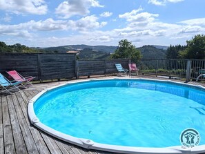 Une piscine ronde, dotée d’une terrasse en bois, entourée d’une clôture métallique, et offrant une vue sur des collines en arrière-plan.
