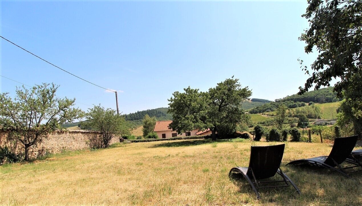 A rural landscape with a stone wall, a building with a red roof, and two lounge chairs in a grassy field.