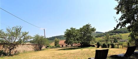 A rural landscape with a stone wall, a building with a red roof, and two lounge chairs in a grassy field.
