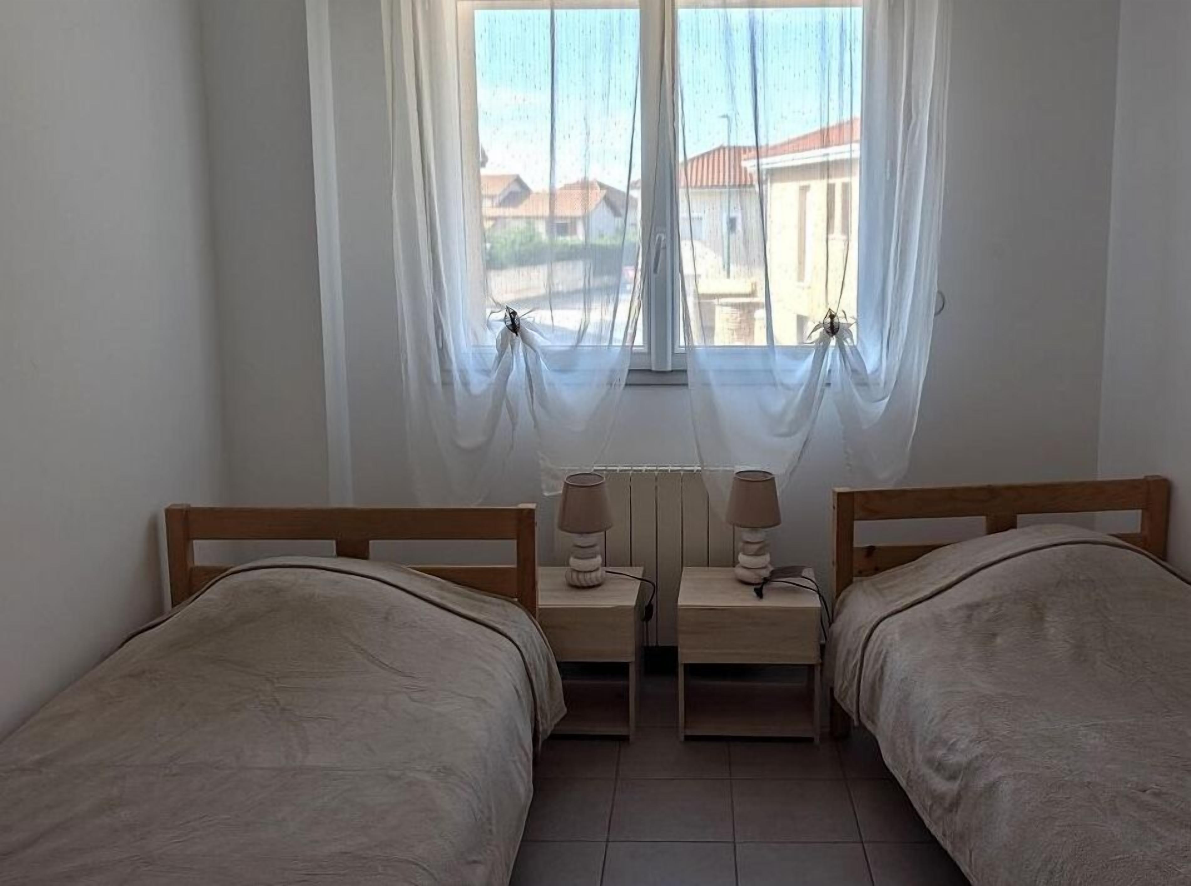 A bedroom with two single beds, a window with sheer curtains, and bedside tables with lamps.