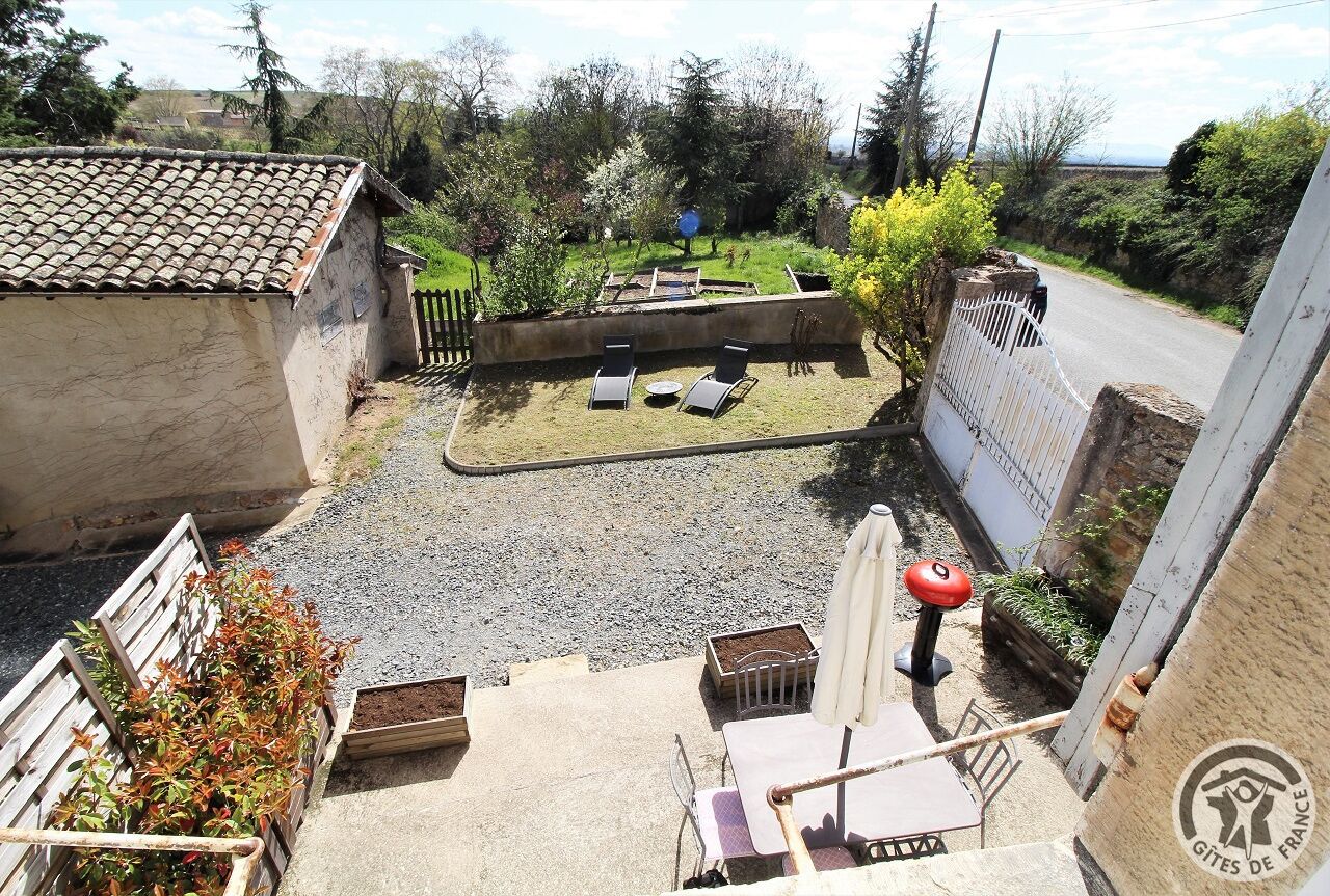 A courtyard with a small seating area, a table, and a chair. There is a white fence surrounding the area, and a gravel path leading to the seating area.