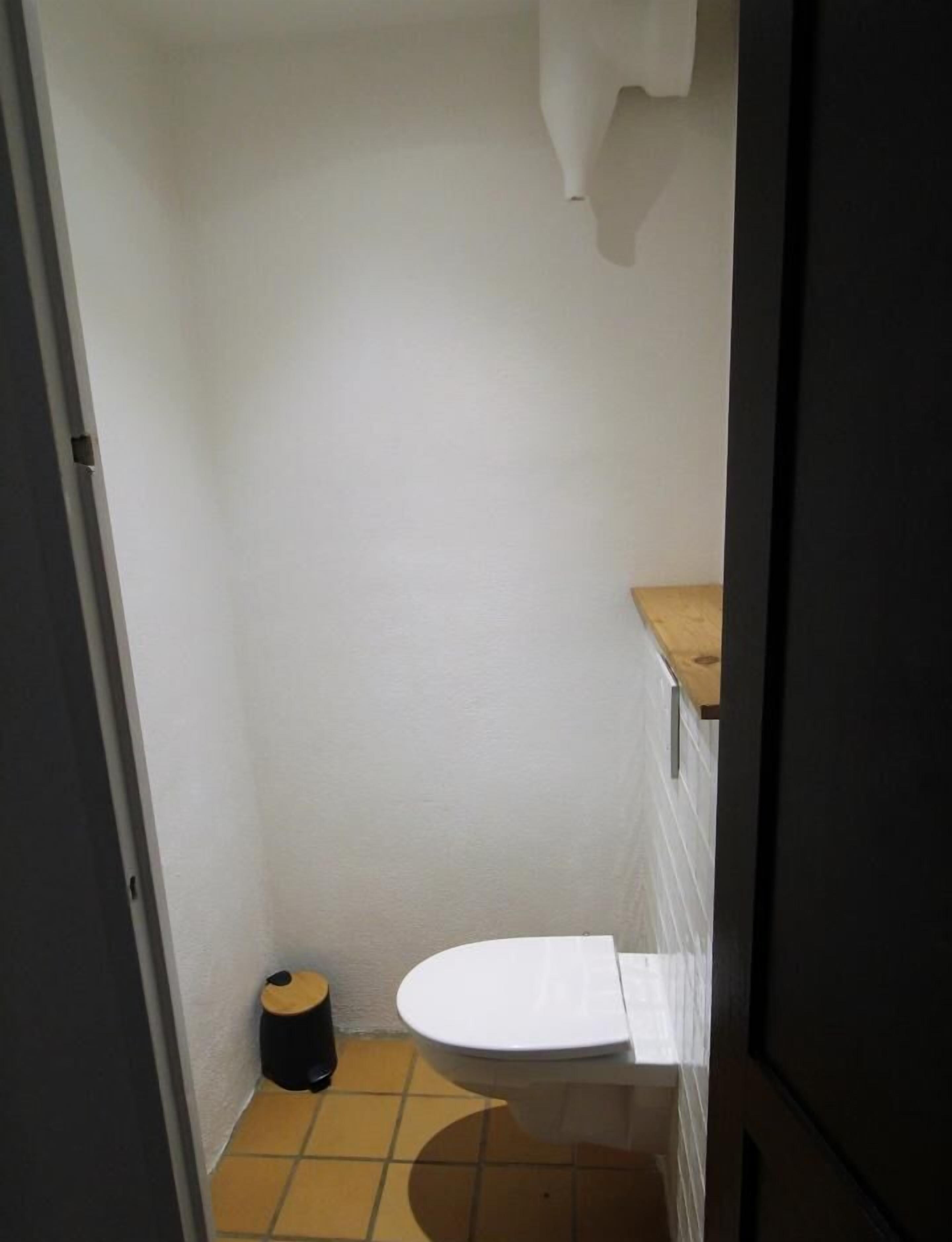 A small, narrow bathroom with a white toilet, a black trash bin, and a wooden shelf above the toilet.
