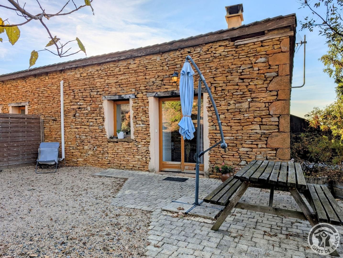 Een stenen gebouw met een houten deur, een klein raam en een terras met een bankje en een tafel.