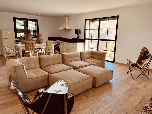 A spacious living room with a sectional sofa, a dining area with wicker furniture, and a kitchen in the background.