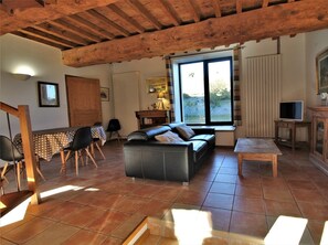 Uma sala de estar espaçosa com sofá de couro preto, mesa de jantar de madeira e cadeiras. Há uma televisão, um abajur e um aparador com espelho.