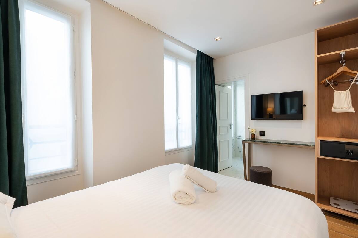A modern hotel room with a large bed, a TV mounted on the wall, a wooden wardrobe, and a desk with a mirror.
