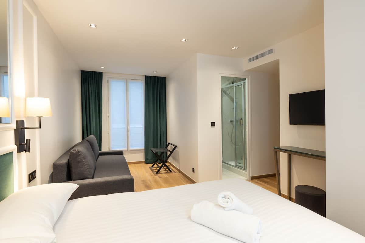 A hotel room with a bed, a sofa, a TV, a glass shower, and a view of the sea.