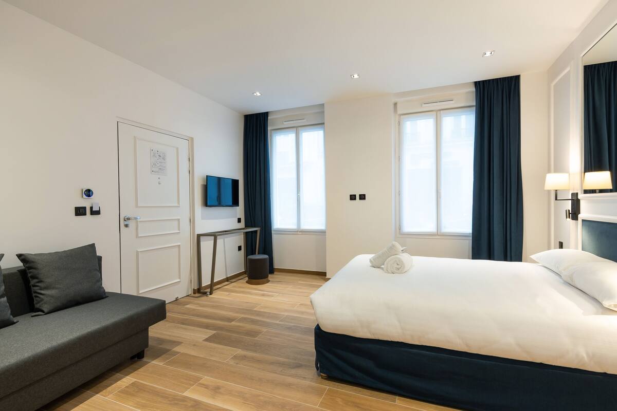 A modern hotel room with a bed, a sofa, a desk, and a TV.