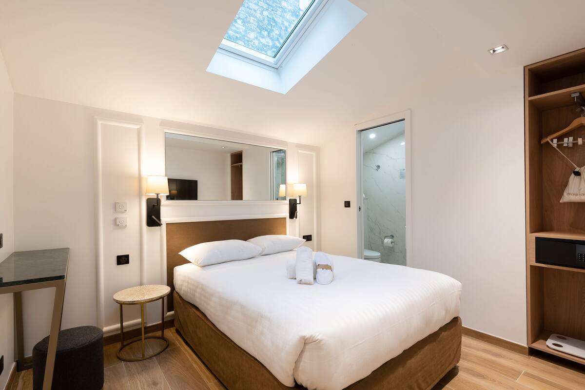 A hotel room with a skylight, a bed with white linens, a bedside table, a mirror, and a wardrobe.