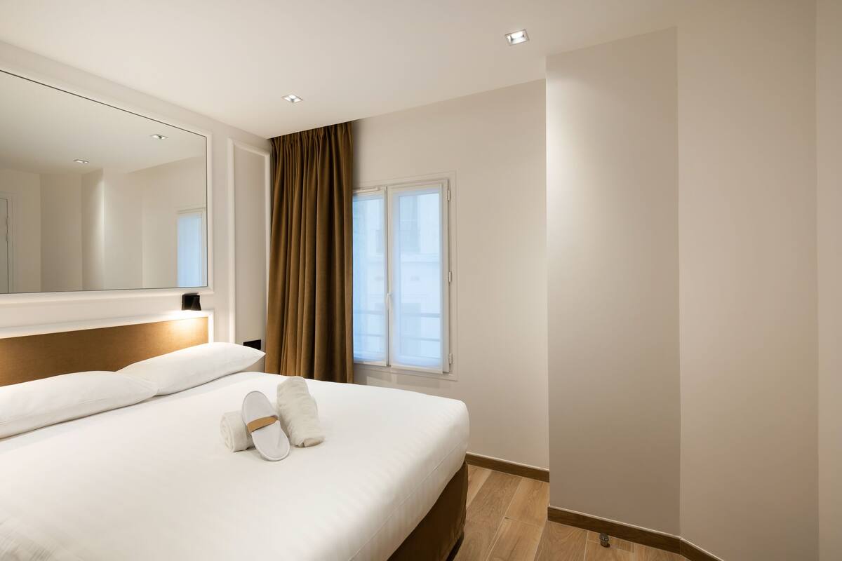 A modern hotel room with a large bed, a mirror, a window with curtains, and a wooden floor.