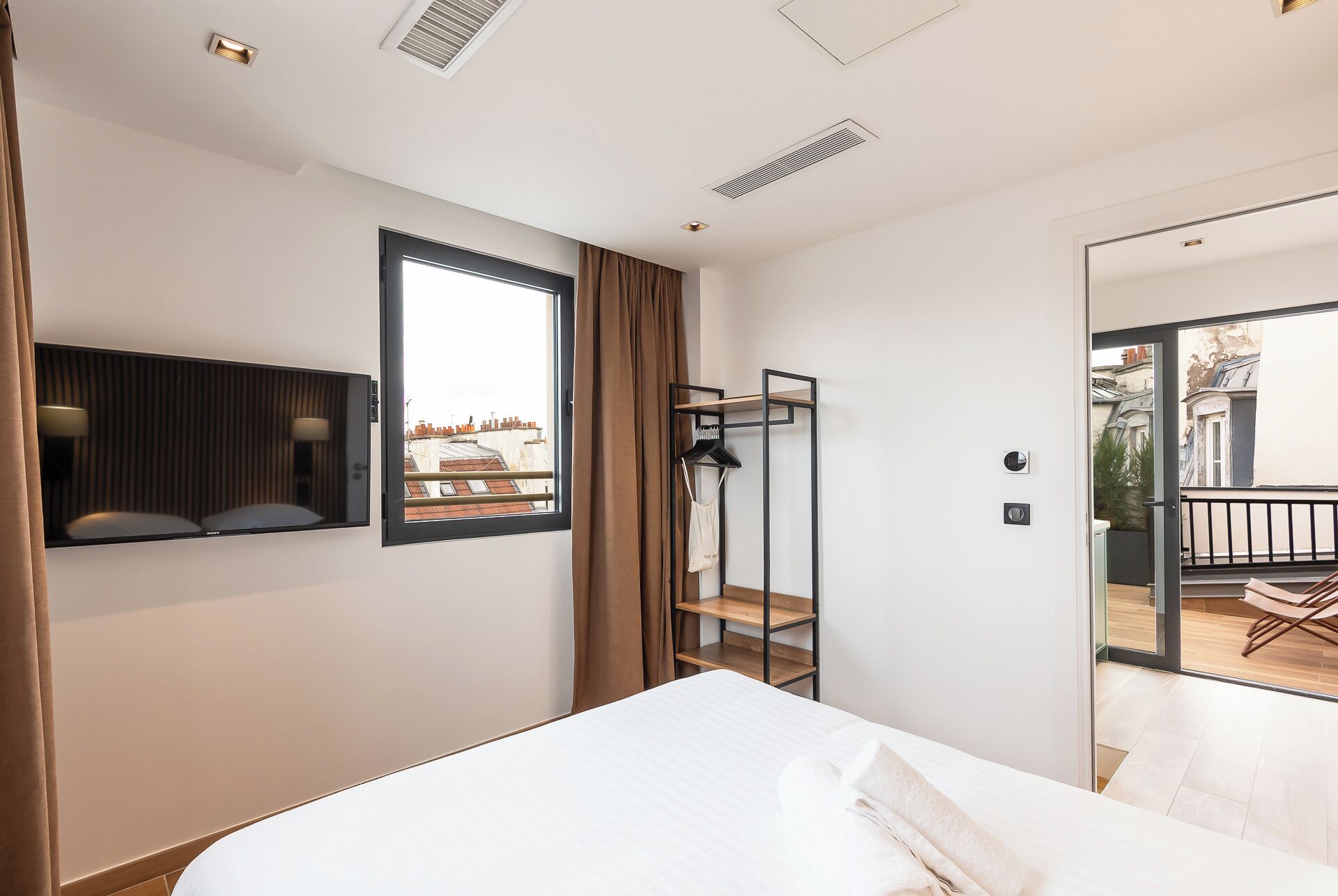 Ein modernes Hotelzimmer mit Bett, Fernseher, Fenster mit Blick auf Gebäude, Balkon mit Tisch und Stühlen sowie Kleiderständer.