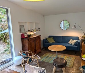 Una sala acogedora con un sofá azul, una mesa de centro redonda de madera, sillones de mimbre, un taburete pequeño y un armario con varios objetos.