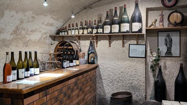 Una bodega con un mostrador de ladrillo que exhibe diversas botellas de vino. Se observan estantes con más botellas y un barril en el suelo.