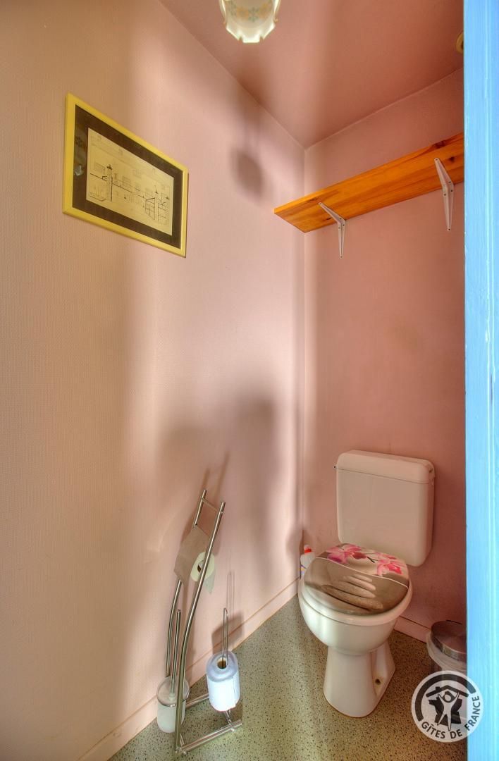 Une petite salle de bain avec des toilettes, une étagère en bois au-dessus, un tableau encadré au mur, et un balai et un seau appuyés contre le mur.