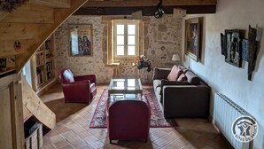 Un salon de style rustique avec un mur en pierre, des poutres en bois au plafond, un canapé en cuir rouge, une table basse en verre et une bibliothèque.