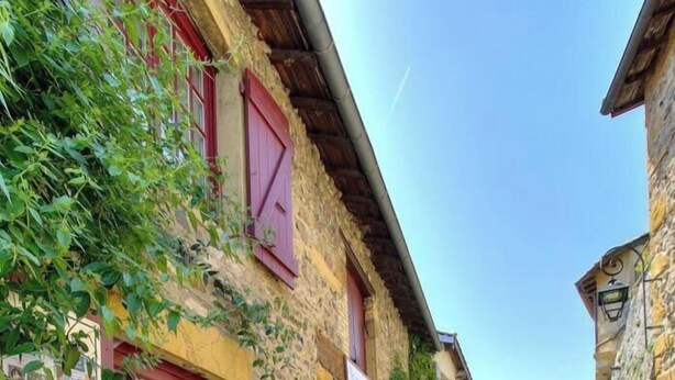Une rue pavée bordée de bâtiments en pierre, avec des volets rouges et un ciel d’un bleu éclatant.