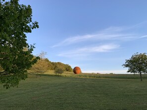 Un campo cubierto de césped con una gran escultura esférica a lo lejos, rodeado de árboles y un cielo despejado.