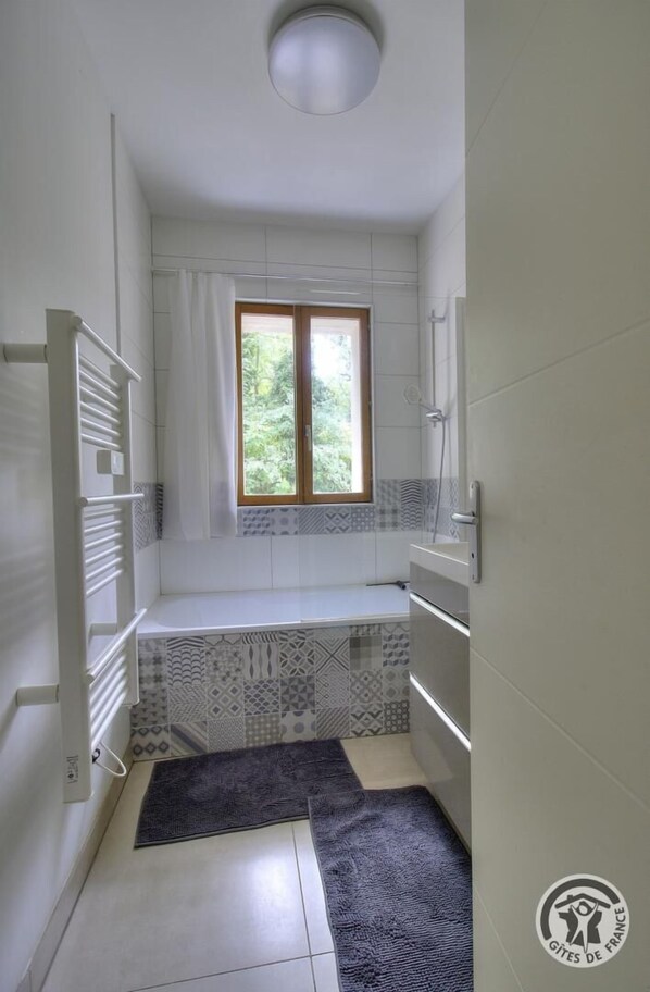 A narrow bathroom with a window, white tiles, a towel rack, and a shower area.