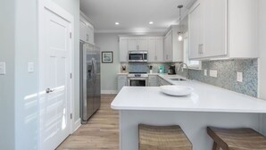 A modern kitchen with white cabinets, a large island, and wooden stools. - The Rookery Iii 6015 3 Bedroom Cottage by RedAwning (Gulf Shores)