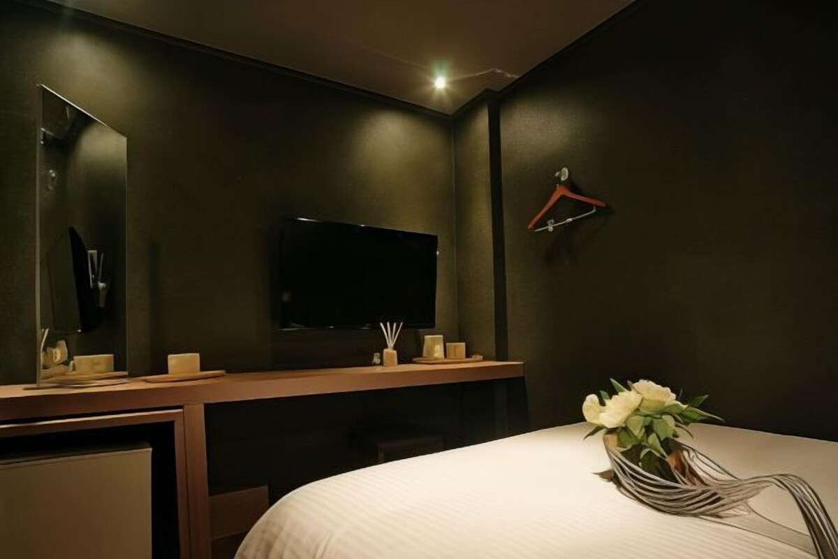 A hotel room with a large bed, a bedside table with a vase of flowers, a flat-screen TV mounted on the wall, and a mirror above the table.