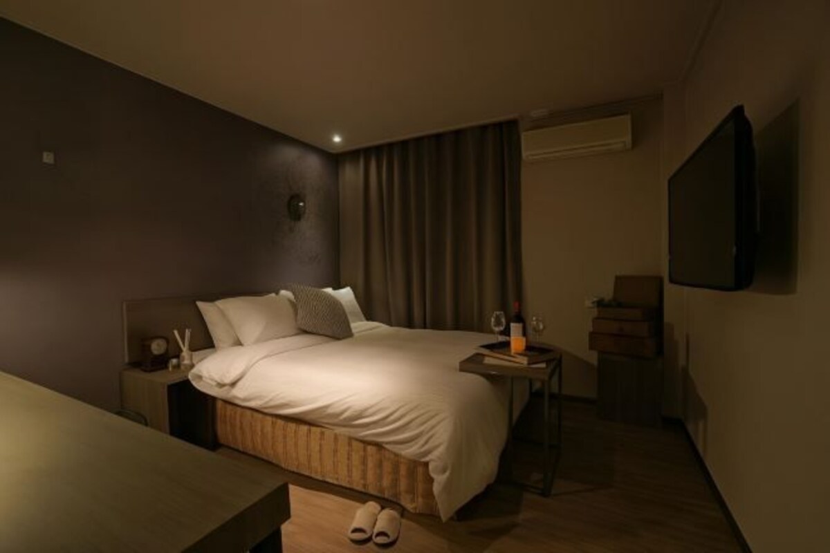 A modern hotel room with a large bed, bedside table with a lamp, a small table with a glass of water, and a TV mounted on the wall.