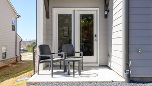 A covered patio with two wicker chairs and a small table, a sliding glass door, and a white exterior wall. - Pet-Friendly 3BR Home Near I-85 and River Trails (Norcross)