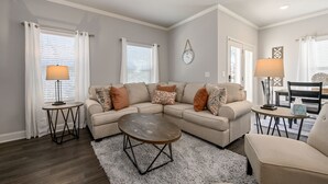 A living room with a beige sectional sofa, a round coffee table, two side tables, and a dining area with a table and chairs. - Pet-Friendly 3BR Home Near I-85 and River Trails (Norcross)