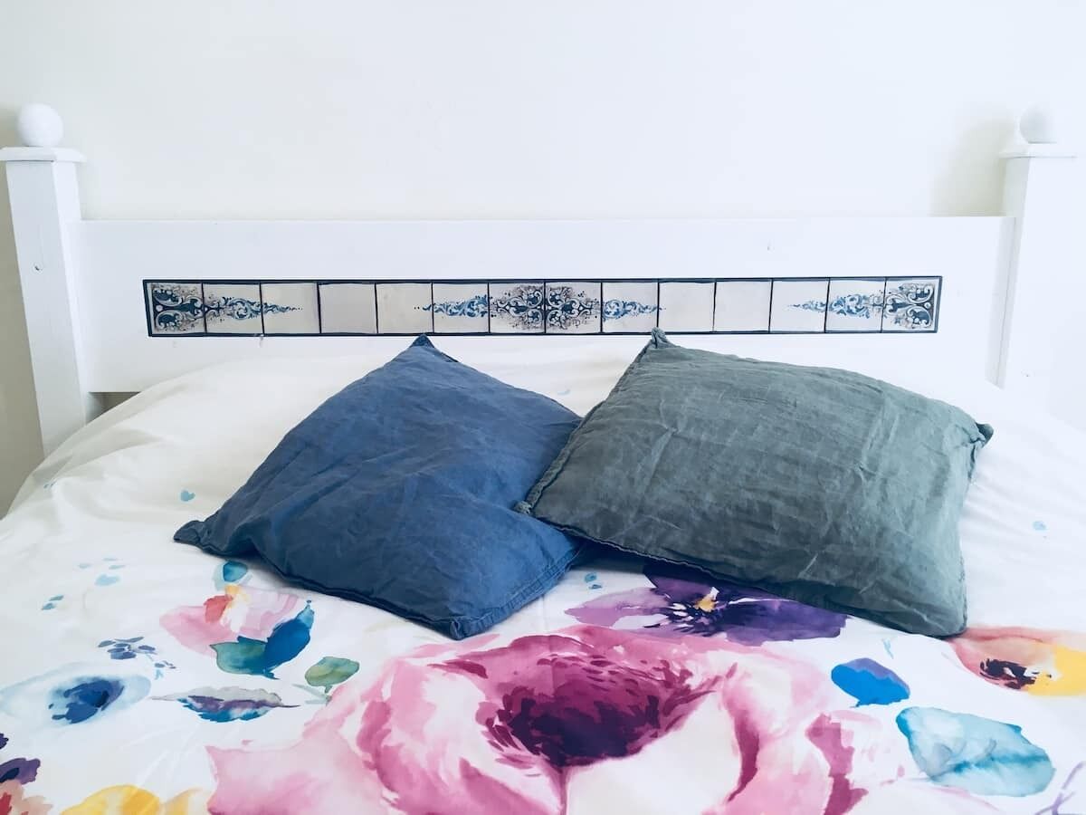 Ein Bett mit einem blumengemusterten Überwurf und zwei Kissen, eines blau und eines grau, an einem weißen Kopfteil mit dekorativen Kacheln.