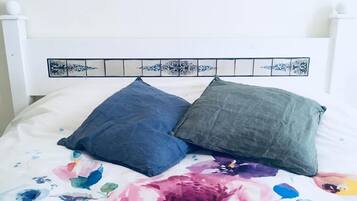 Ein Bett mit einem blumengemusterten Überwurf und zwei Kissen, eines blau und eines grau, an einem weißen Kopfteil mit dekorativen Kacheln.