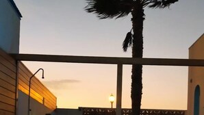 A swimming pool with a palm tree and lounge chairs, surrounded by buildings with a sunset in the background. - Cozy Villa by MAHBO (A) (Corralejo)