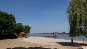 A sandy beach with two wooden structures, one with a red roof and another with a brown roof, near a calm body of water. - Mikin dol Baranja Country House (Draz)