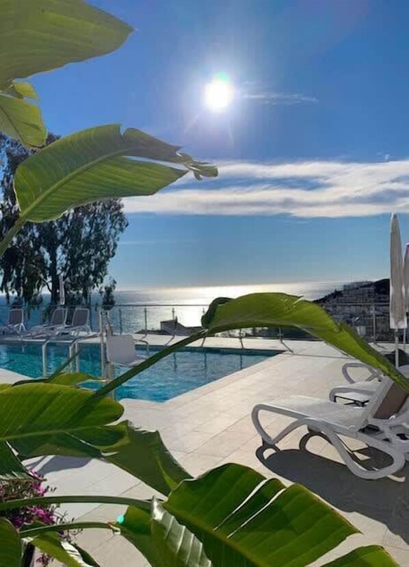 A poolside area with lounge chairs, a view of the sea, and tropical plants.