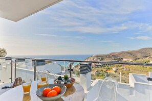 A balcony with a table, a bowl of fruit, a glass of juice, and a view of the sea and hills.