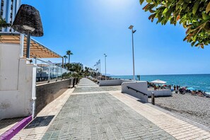 A seaside promenade with a tiled walkway, palm trees, and beach umbrellas.